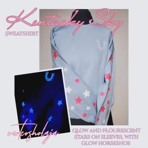 May include: Light blue sweatshirt with pink and white stars on the sleeves and a white horseshoe design. The text "Kentucky Sky" and "Sweatshirt" are in pink script. The image also includes a glowing image of stars and a horseshoe.
