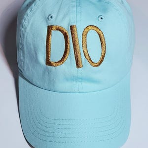 Op de afbeelding: Lichtblauwe baseballcap met de geborduurde letters "DIO" in goud. De cap heeft een gebogen klep en een ontwerp met zes panelen met ventilatiegaten.
