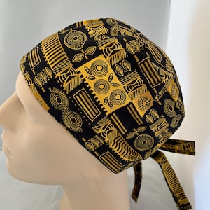 May include: A black and gold patterned scrub cap with a tie closure. The cap features a repeating geometric design in gold against a black background. The cap is shown on a mannequin head.