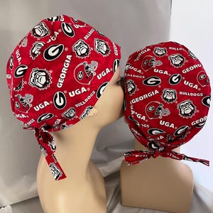 May include: Two red scrub caps with a repeating pattern of white bulldog logos, football helmets, and the text "UGA" and "GEORGIA". The caps are tied at the back with matching red fabric.