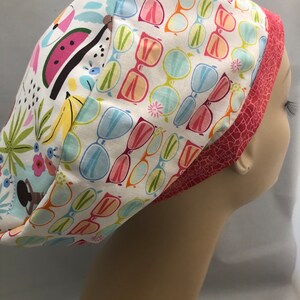 Puede incluir: Un gorro de quirófano blanco con un patrón colorido de gafas de sol, plátanos, sandía y flores. El gorro tiene un ribete rosa y rojo.