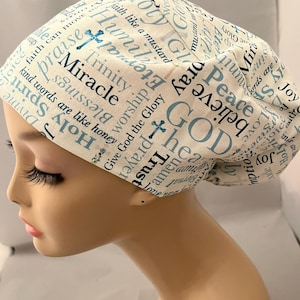 May include: A white and light blue patterned scrub cap featuring religious words like "Miracle", "God", and "Faith". The cap has a cross design and is made of a soft fabric.