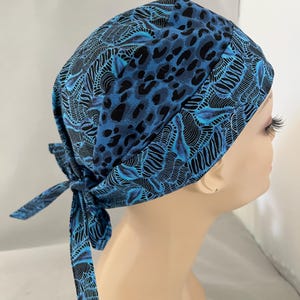 May include: A blue and black patterned scrub cap with a tie closure. The cap features a mix of leopard print and abstract designs. The cap is tied at the back, and is shown on a mannequin head.