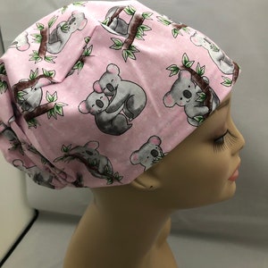 May include: Pink surgical scrub hat with a repeating pattern of grey koalas on branches. The koalas are in various poses, including hugging.