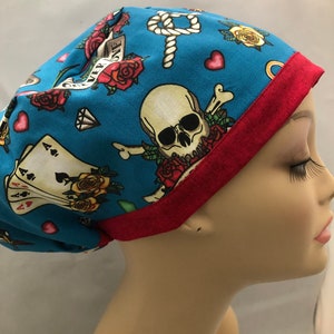 May include: A blue scrub hat with a red trim. The hat has a skull and crossbones design with roses, hearts, playing cards, and a rope knot.