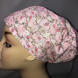 May include: A pink and white scrub hat with a pattern of pink pigs on a white background. The pigs are cartoon-like and have a playful expression.