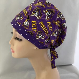 May include: Purple and gold surgical scrub hat with a white polka dot trim. The hat features a repeated pattern of the East Carolina University Pirates logo, including the words "ECU Pirates" and "East Carolina".