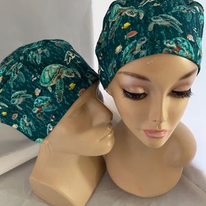 May include: Two teal scrub caps with a sea turtle and fish pattern. The caps are on mannequin heads. The fabric has a dark teal background with illustrations of sea turtles, yellow and orange fish, and bubbles.