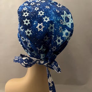 May include: A blue surgical scrub cap featuring a celestial design with white stars and a scattering of white dots. The cap is tied at the back with a matching fabric bow, showcasing a festive pattern of menorahs and other holiday symbols.