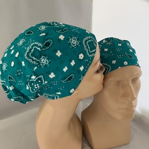 May include: Two teal paisley patterned scrub caps on mannequin heads. The caps feature a repeating pattern of white and black paisley designs. One cap is on a head with a face, and the other is on a head without a face.
