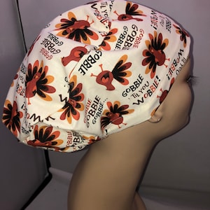 May include: A white Thanksgiving-themed bouffant surgical scrub cap. The cap features a repeating pattern of cartoon turkeys in shades of brown, orange, and black, along with the words "GOBBLE 'Til You WOBBLE!" in black.