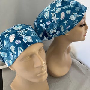 May include: Two scrub caps featuring a deep teal background with a repeating pattern of white and light blue seashells, starfish, coral, and sand dollars. The caps are designed with a rounded crown and a wide band that fits around the head.