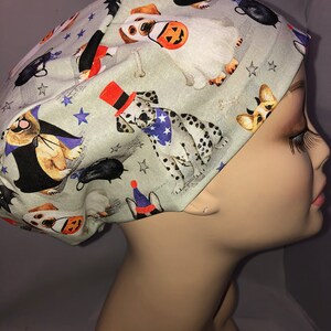 May include: A light gray surgical scrub cap with a Halloween-themed dog print. The pattern includes dogs dressed as ghosts, vampires, and wearing hats, along with stars and other festive elements. The cap is designed to fit comfortably.