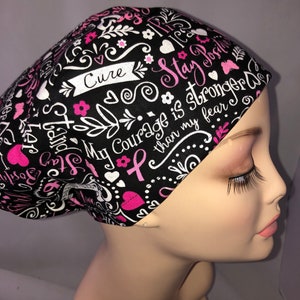 May include: Black scrub hat with a pink and white design. The hat features a pink ribbon, hearts, and the text "Stay Strong, My Courage is Stronger than my Fear, Cure".