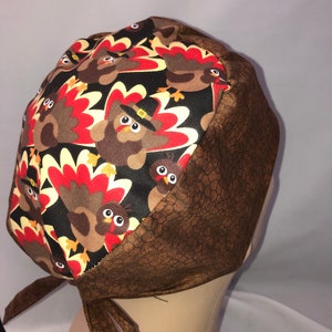 Pode incluir: Uma touca cirúrgica com tema de Ação de Graças com um padrão repetido de desenhos animados de perus com penas vermelhas, amarelas e marrons em um fundo preto. A touca tem uma borda marrom texturizada e laços.
