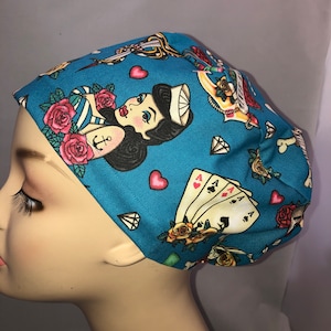 May include: A blue scrub hat with a colorful print of a pin-up girl, roses, playing cards, and other tattoo-style imagery.