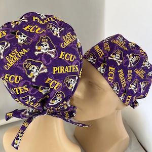 May include: Two purple scrub caps with a repeating pattern of pirate skull and crossbones, the words "ECU" and "East Carolina Pirates" in yellow. One cap has a bow tie. The caps are displayed on mannequin heads.