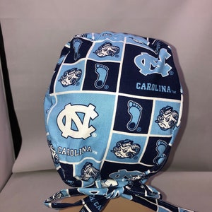 Pu&ograve; includere: Un berretto chirurgico blu e bianco con motivo con il logo degli UNC Tar Heels e il testo "CAROLINA TM".