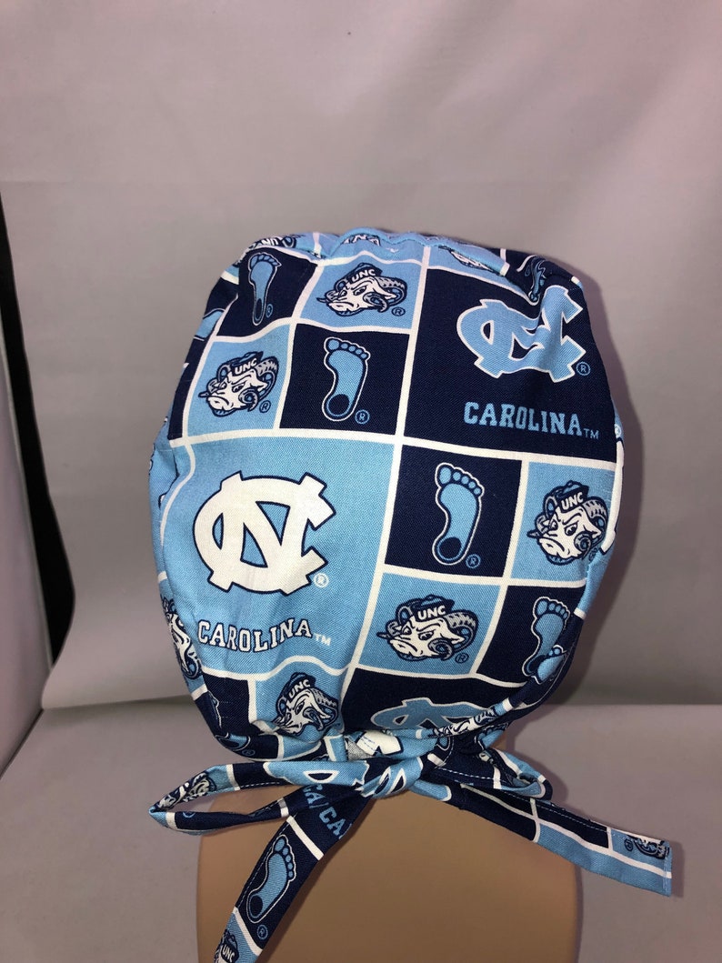 Pu&ograve; includere: Un berretto chirurgico blu e bianco con motivo con il logo degli UNC Tar Heels e il testo "CAROLINA TM".