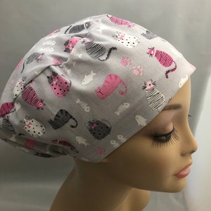 May include: A gray surgical scrub cap featuring a repeating pattern of stylized cats in pink, black, and white. The cats are interspersed with fish bones and small pink floral accents. The cap is designed to cover the head.
