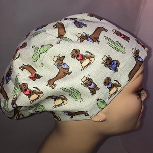 May include: A light green scrub hat with a pattern of brown dachshunds wearing cowboy hats and bandanas. The dachshunds are surrounded by green cacti.