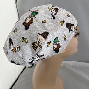 May include: A gray scrub cap featuring a repeating pattern of cartoon characters in various poses. The characters are depicted in black, brown, yellow, and white. The cap is made of a soft fabric and is designed to cover the head.