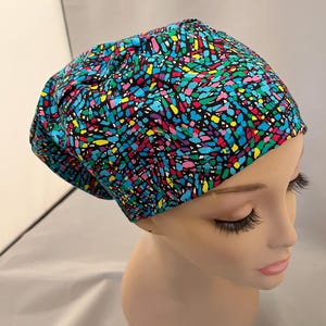 May include: A colorful scrub cap with a vibrant pattern of blue, green, yellow, pink, and purple shapes on a black background. The cap is worn by a mannequin head, showcasing its design and fit.