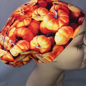 May include: A patterned scrub cap featuring a repeating design of orange pumpkins. The cap is made of fabric and is worn on a mannequin head. The pumpkins have green stems and are set against a dark background.