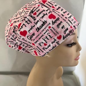 May include: A pink scrub cap with a repeating pattern of red hearts and inspirational phrases in black script. The cap is designed to be worn in a medical or caregiving setting, offering a touch of positivity.