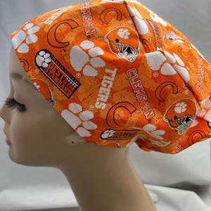 May include: Orange and white patterned scrub cap featuring Clemson Tigers football team logos, paw prints, and text. The cap is made of a soft fabric and is designed to be worn in a medical or professional setting.