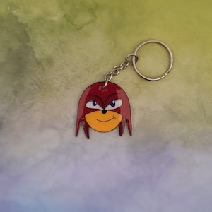 May include: Red and yellow cartoon character keychain with a silver ring. The character has a red head with a yellow face and blue eyes.
