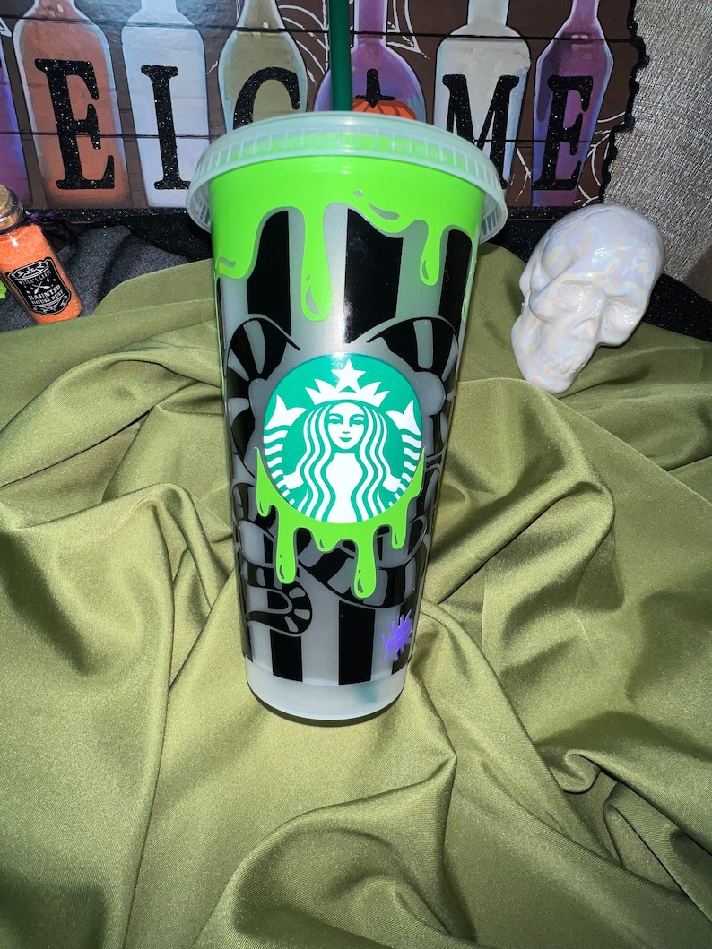 Pode incluir: Um copo reutiliz&aacute;vel verde e preto com um logotipo da Starbucks e um design de slime verde. O copo tem uma tampa transparente e um canudo.