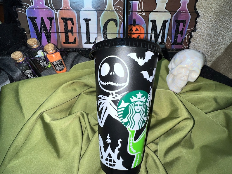 Puede incluir: Taza reutilizable negra con un dise&ntilde;o blanco que presenta al personaje Jack Skellington de la pel&iacute;cula Pesadilla antes de Navidad, un logotipo de Starbucks y murci&eacute;lagos. La taza tiene una tapa verde y una pajita.