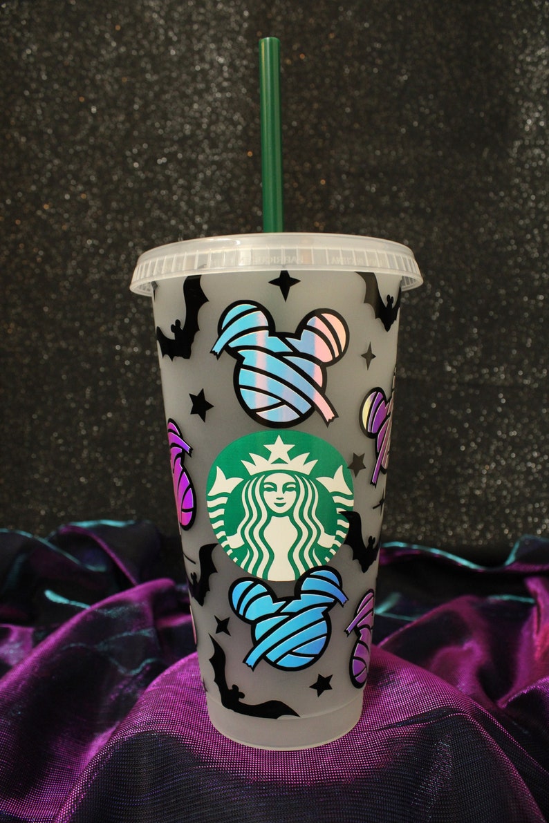 Peut inclure: Un gobelet Starbucks en plastique transparent avec une paille et un couvercle verts. La tasse est d&eacute;cor&eacute;e de chauves-souris noires, d'&eacute;toiles et de t&ecirc;tes de Mickey Mouse bleues, roses et violettes. Un logo Starbucks vert est centr&eacute; sur la tasse.