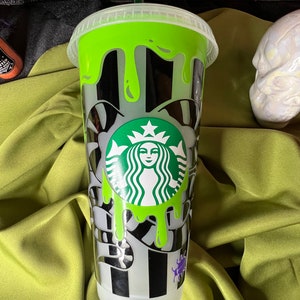 Pode incluir: Um copo reutiliz&aacute;vel de pl&aacute;stico transparente com um design listrado preto e branco e slime verde pingando pelo lado. O copo tem um logotipo verde Starbucks com o rosto de uma mulher no centro.