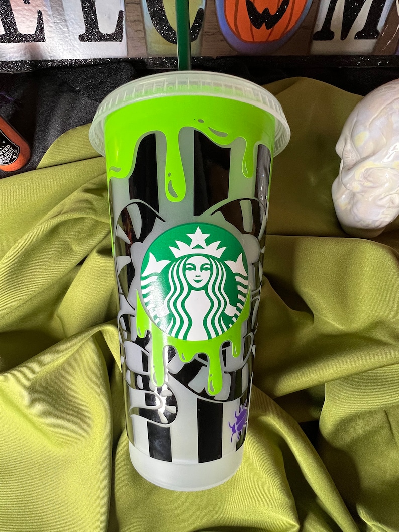 Pode incluir: Um copo reutiliz&aacute;vel de pl&aacute;stico transparente com um design listrado preto e branco e slime verde pingando pelo lado. O copo tem um logotipo verde Starbucks com o rosto de uma mulher no centro.
