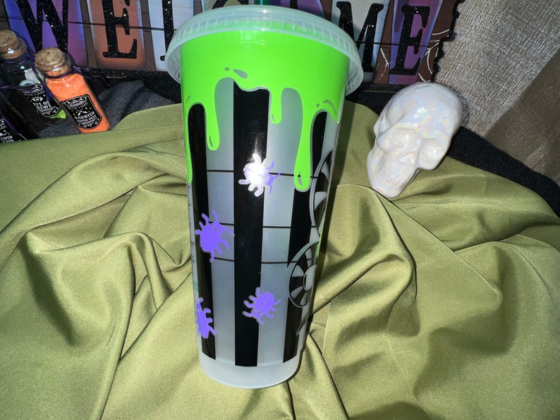 Pode incluir: Um copo reutiliz&aacute;vel com listras pretas e brancas com um design de slime verde e detalhes de aranha roxa. O copo tem uma tampa transparente.