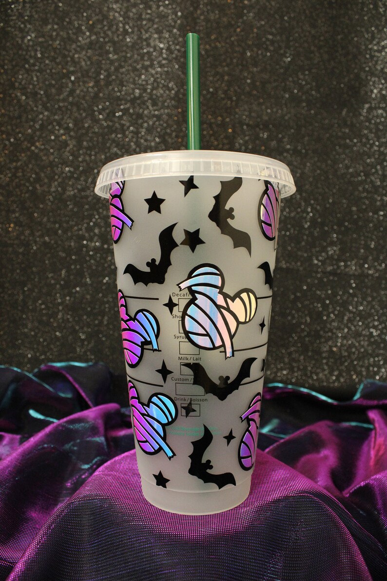 Peut inclure: Un gobelet en plastique transparent avec un couvercle et une paille verte. Le gobelet est d&eacute;cor&eacute; de chauves-souris noires, d'&eacute;toiles et de motifs de Mickey Mouse sur le th&egrave;me de la momie aux couleurs iris&eacute;es. Le gobelet comporte des rep&egrave;res de mesure.