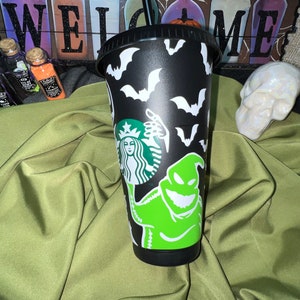 Puede incluir: Taza reutilizable negra con un personaje Oogie Boogie verde y blanco de Pesadilla antes de Navidad, un logotipo de Starbucks y murci&eacute;lagos.