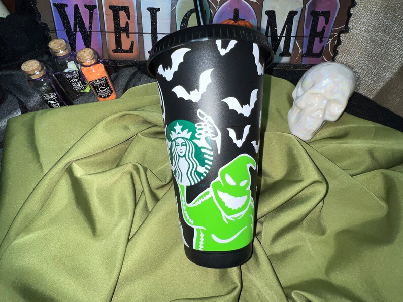 Puede incluir: Taza reutilizable negra con un personaje Oogie Boogie verde y blanco de Pesadilla antes de Navidad, un logotipo de Starbucks y murci&eacute;lagos.