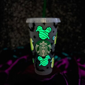 Peut inclure: Un gobelet froid Starbucks en plastique transparent avec une paille verte. La tasse est d&eacute;cor&eacute;e d'images sur le th&egrave;me d'Halloween qui brillent dans le noir, notamment des chauves-souris, des &eacute;toiles et des t&ecirc;tes de Mickey Mouse envelopp&eacute;es. Le logo Starbucks est visible.
