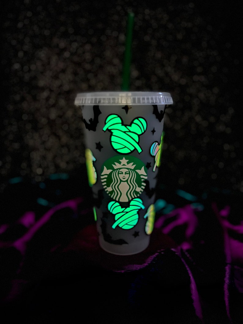 Peut inclure: Un gobelet froid Starbucks en plastique transparent avec une paille verte. La tasse est d&eacute;cor&eacute;e d'images sur le th&egrave;me d'Halloween qui brillent dans le noir, notamment des chauves-souris, des &eacute;toiles et des t&ecirc;tes de Mickey Mouse envelopp&eacute;es. Le logo Starbucks est visible.
