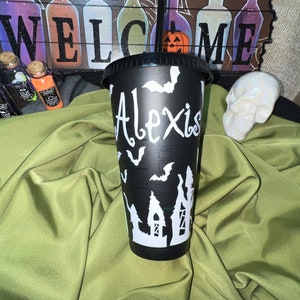 Puede incluir: Vaso reutilizable negro con un dise&ntilde;o de Halloween blanco con murci&eacute;lagos, una casa encantada y el nombre "Alexis".