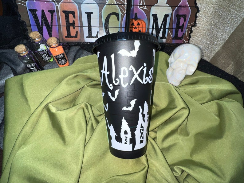 Puede incluir: Vaso reutilizable negro con un dise&ntilde;o de Halloween blanco con murci&eacute;lagos, una casa encantada y el nombre "Alexis".