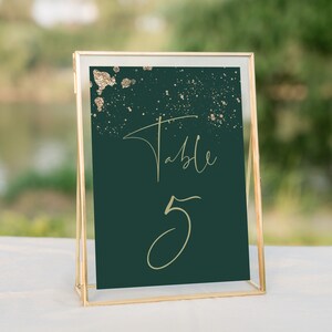 May include: A gold-framed table number sign with a dark green background and gold glitter accents. The sign reads "Table 5" in gold script.