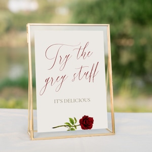 Puede incluir: Un cartel con marco dorado con el texto "Try the grey stuff. It's delicious." en escritura color burdeos. Una sola rosa roja con hojas verdes se encuentra en la parte inferior del cartel.