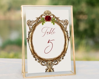 May include: A gold-framed table number card with a red rose and green leaves. The card reads "Table 5" in a cursive font.