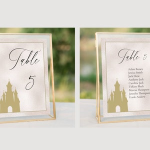 K&ouml;nnte beinhalten: Zwei goldgerahmte Tischnummernschilder mit einer Schlosssilhouette und dem Text "Tisch 5". Das Schild rechts listet auch die Namen der G&auml;ste auf, die dem Tisch zugewiesen sind.