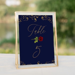 May include: A gold-framed table number sign with a navy blue background. The sign features a gold-colored floral design, a red rose, and the text "Table 5" in gold.