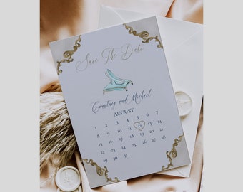 May include: A light blue save the date card with a gold ornate border. The card features a watercolor illustration of a glass slipper and the text "Save the Date" in cursive. The text "Corrtrey and Michael" is printed below the illustration, followed by the month "AUGUST" and a calendar with the date "12" circled in a heart.
